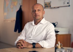 A confident adult man in a white shirt sitting in a well-lit office with a thoughtful expression.